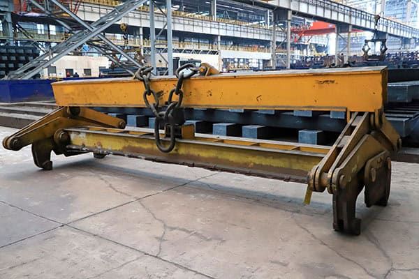 Industrial Yellow Spreader Beam in Factory Workshop A close-up shot of a heavy-duty yellow industrial spreader beam with black chain slings and hooks, securely placed on a concrete floor in a modern factory workshop. The background features clear industrial steel structures, blue safety barriers, and neatly arranged metal raw materials, showcasing the rigor and bustle of heavy industry production.